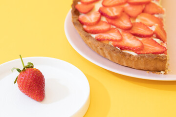 Piece of grated with strawberries and cream on a pink plate. Yellow background. Strawberry on a white plate in the foreground. - 435213507