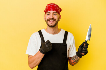 Young tattooed batcher caucasian man holding a knife isolated on yellow background smiling and raising thumb up