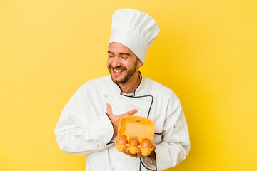 Young caucasian chef man holding eggs isolated on yellow background laughs out loudly keeping hand on chest.
