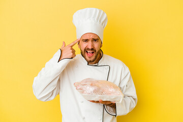 Young caucasian chef man holding chicken isolated on yellow background showing a disappointment gesture with forefinger.