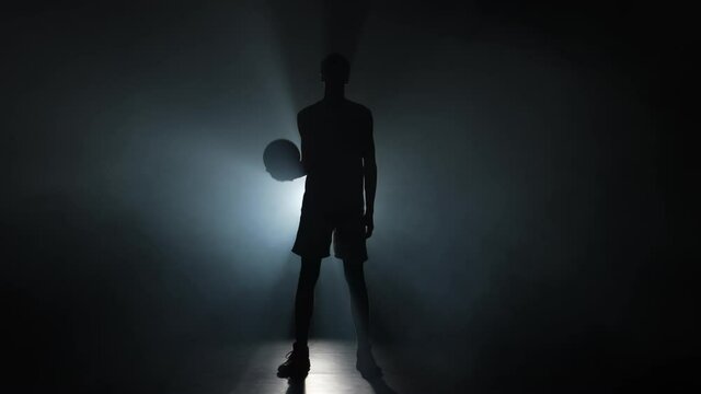 Silhouette of a young guy basketball player tossing the ball up. Tall young sportsman in sports uniform posing on a dark studio background with smoke and backlight. Slow motion ready 59.94fps.