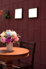 Side view of artificial rose flowers in white ceramic vase on wooden round table with teak wood chair and blank picture frame on wooden wall background in vintage style and vertical frame