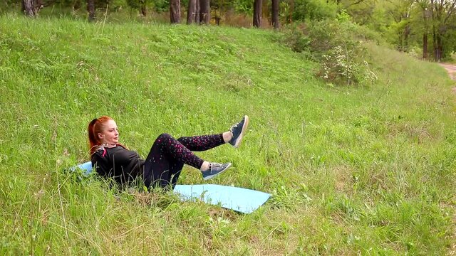 40-year-old beautiful woman with bright red hair works out in the park, does exercises for a tummy tuck and listens to music in wireless headphones