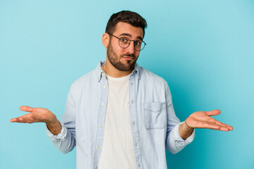 Young caucasian man isolated on blue background doubting and shrugging shoulders in questioning gesture. - obrazy, fototapety, plakaty