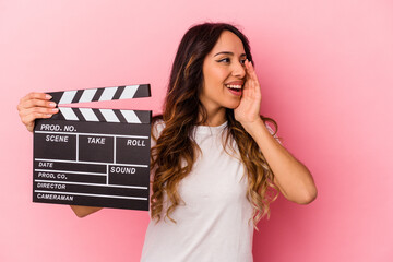 Young mexican woman holding clapperboard isolated on pink background shouting and holding palm near...