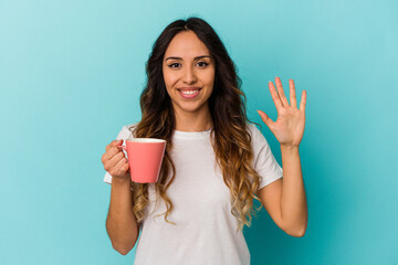 Young mexican woman holding a mug isolated on blue background smiling cheerful showing number five with fingers.