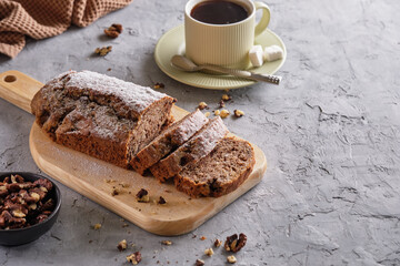 Delicious and flavorful banana cake with nuts and chocolate on a wooden board with a cup of coffee on a gray background