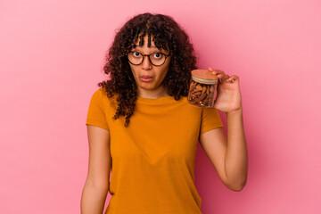 Young mixed race woman holding an almond jar isolated on pink background shrugs shoulders and open...