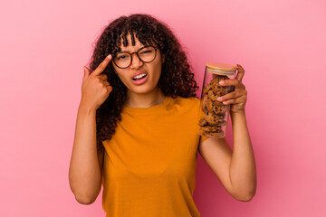 Young mixed race woman holding a cookies jar isolated on pink background showing a disappointment gesture with forefinger.