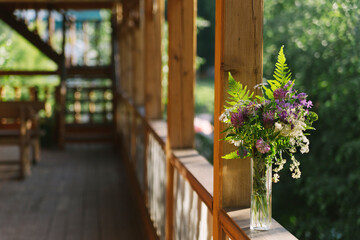 A summer field bouquet in a small vase stands on the veranda of a large wooden house or an old-style summer cottage.
