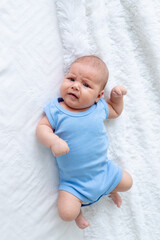 crying baby boy three months old in a blue bodysuit on a white bed at home