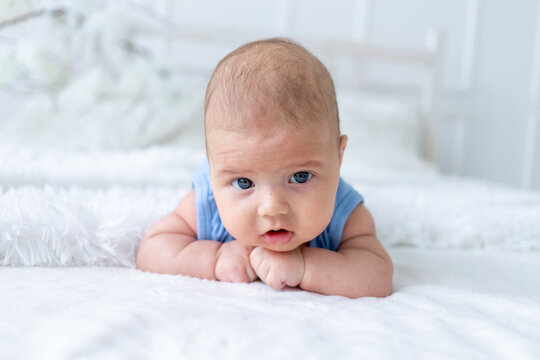 cute baby boy three months old in a blue bodysuit lies on his stomach on a white bed at home, the newborn learns to hold his head