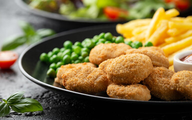 Fried crispy chicken nuggets with ketchup, french fries and green peas in black plate