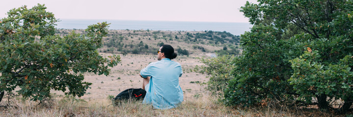 A male tourist with a backpack sits on the top of the hill.