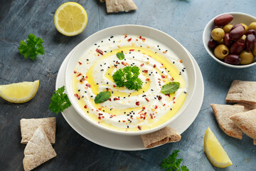 Labneh cream cheese dip with olive oil, salt, herbs served with olives, pita bread in white bowl