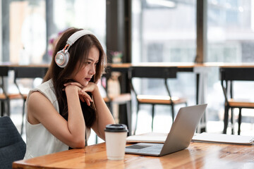 Obraz premium Concentrated young asian female wearing headphone look at laptop for online study, attractive female student learning language, watching online webinar, e-learning education concept.