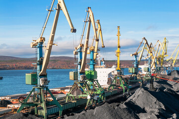 Heaps of coal in the Murmansk Commercial Sea Port. Loading of coal by buckets (grabs) of portal cranes into the holds of a dry cargo ship in the seaport
