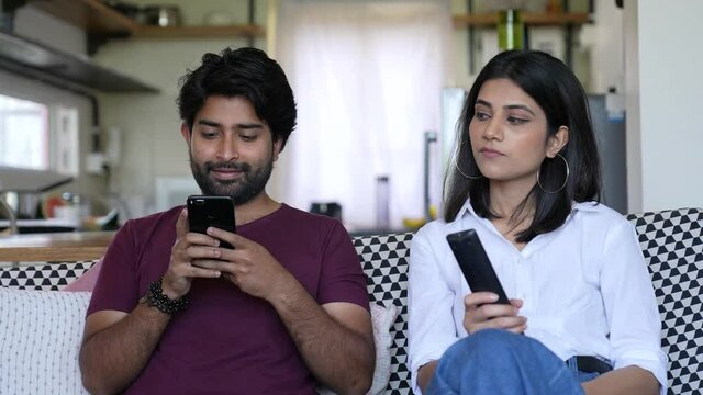 Smiling Asian Man Using Smart phone do chat  and woman looking him while sitting at couch at home indoor shot.lifestyle concept.social distance.Asian woman watching television while looking him.