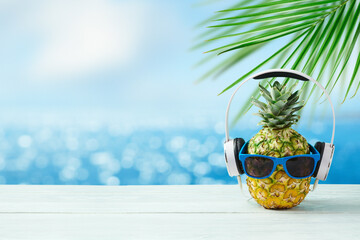 Pineapple with headphones and sunglasses on wooden table over bokeh sea beach background. Summer...