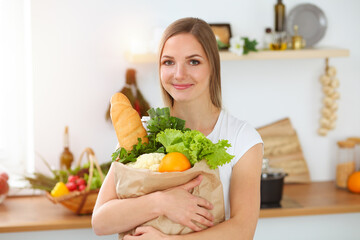 Obraz na płótnie Canvas An attractive young woman holding the paper bag full of vegetables while standing and smiling in sunny kitchen. Cooking concept