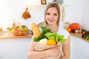 An attractive young woman holding the paper bag full of vegetables while standing and smiling in sunny kitchen. Cooking concept