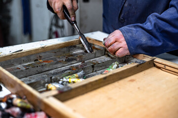 Man cleaning an old paint set briefcase by scraping it with a chisel in his hand. Tidying and neatening an used oil paint kit during a painting workshop activity in a studio. Work of a painter’s trade