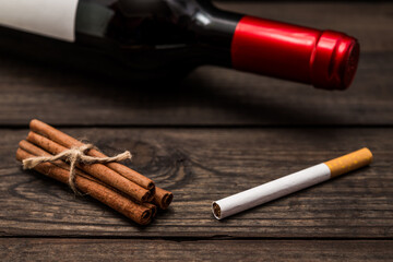 Bottle of red wine and cinnamon sticks tied with jute rope with cigarette lying on an old wooden table. Close up view, focus on the cigarette