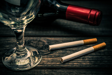 Bottle of red wine and two cigarettes with empty wineglass on an old wooden table. Close up view, focus on the cigarette