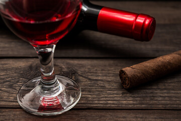 Bottle of red wine with a glass of red wine and cuban cigar on an old wooden table. Close up view, focus on the cuban cigar
