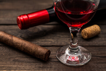 Bottle of red wine with a glass of red wine and cuban cigar on an old wooden table. Close up view, focus on the cuban cigar