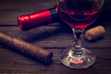 Bottle of red wine with a glass of red wine and cuban cigar on an old wooden table. Close up view, focus on the cuban cigar