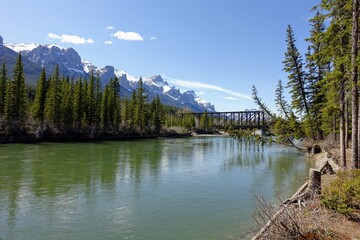 A beautiful photo of the bow river in Canmore, Alberta, on a beautiful sunny spring day, with the rocky mountains and walking bridge in the background. The view is from a walking path along the river