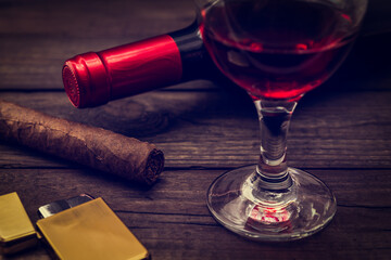 Bottle of red wine with a glass of red wine and cuban cigar with golden lighter on an old wooden table. Close up view, focus on the cuban cigar