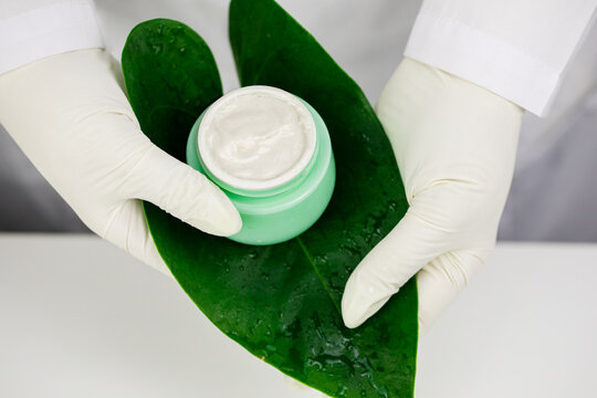 gloved hands of female lab technician hold Luxury skincare moisturizing cream in open green glass jar on large tropical plant leaf close up top view