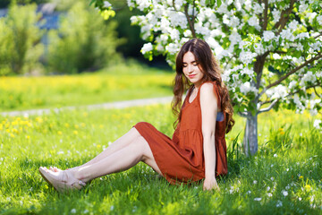 Naklejka na ściany i meble A young woman is sitting under a blooming apple tree against a blurred background. Shallow focus.