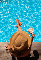 A pretty little girl sits in a hat by the pool and holds a cup of cocoa and marshmallows in her hand. Beautiful close-up hat. A long-awaited vacation. Clean swimming pool.