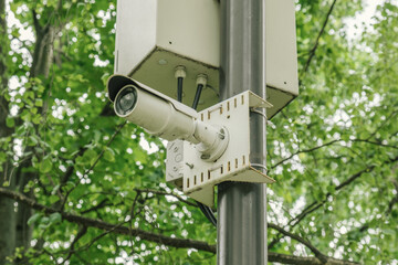 Small white CCTV camera on on a lamppost in a city park against a green tree branches. Security, video surveillance, face recognition.