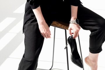 Young woman in black business suit sits on the stool, holding high-heeled shoe in her hand