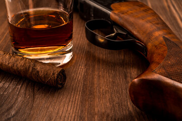 Hunting rifle and glass of whiskey with cuban cigar on the wooden table. Focus on the cuban cigar