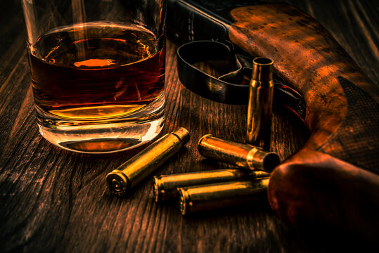 Empty shells and the rifle with glass of whiskey on a wooden table. Focus on the glass of whiskey