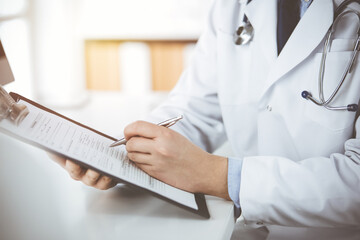 Unknown male doctor and patient woman discussing current health examination while sitting in sunny clinic and using clipboard. Perfect medical service in hospital