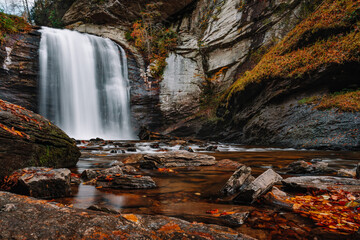 A plunge pool waterfall known as Looking Glass Falls surrounded by logs, stones and colorful fall foliage in Pisgah National Forest, Transylvania County 