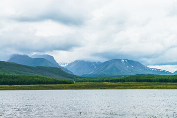 Dramatic landscape with mountain lake and forest on hills in sunlight and snowy mountains in low clouds in changeable weather. High mountains in cloudy sky and lake near green grasses and forest hills