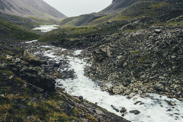 Atmospheric landscape with mountain lake and mountain creek among moraines in rainy weather. Bleak overcast scenery with milky river and lake among rocks. Gloomy view to milk mountain river and lake.