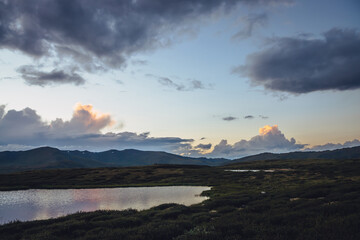 Scenic mountain scenery with lilac reflection of sunset in lake. Colorful mountain landscape with illuminating color in violet clouds in dawn gradient sky. Beautiful view to sunrise and lilac lake.