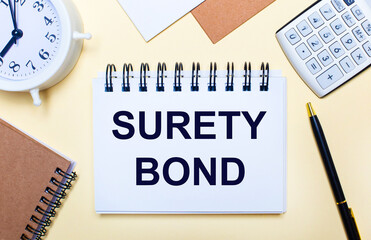 On a light background, a white alarm clock, a calculator, a pen and a notebook with the text SURETY BOND. Flat lay