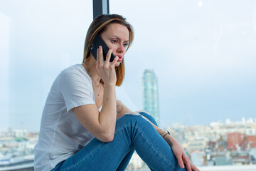 Sad girl in a white T-shirt talking on the phone sitting on the windowsill with panoramic windows overlooking the city. High quality photo
