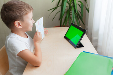 Smiling school boy siting at desk and looks at tablet with green screen. Happy child showing his...