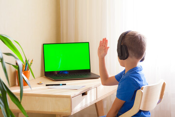 smart school boy in headphones sits at the table and looks at the computer. A child pulls a hand to answer. Online lesson at a distance learning. Green screen chroma key on the monitor.