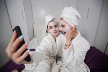 Gay young women take selfies in face masks and white robes, have fun at home. Beauty and personal care concept. High quality photo